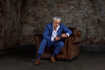 A handsome middle-aged man in a blue formal suit poses sitting on a leather armchair. Portrait of a businessman. A handsome middle-aged man poses on a brick background with a place to copy