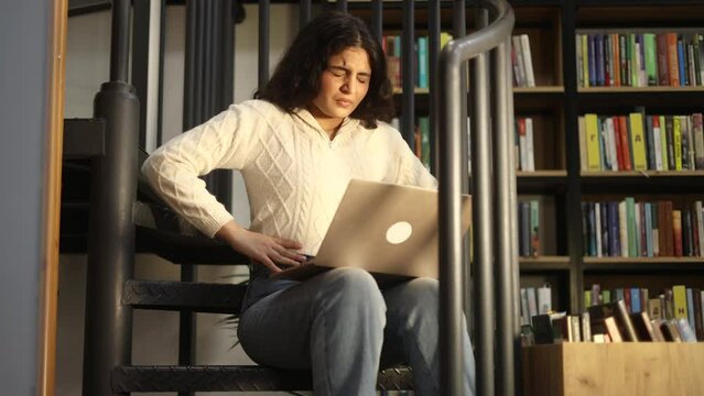 Sick unhealthy young curly woman student clasping belly and suffering from severe abdominal pain periods spasm irritable bowel syndrome of diet at university library workplace