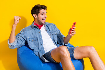 Portrait of overjoyed man dressed denim shirt red scarf sit on bean bag look at smartphone win...