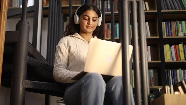 Portrait of pretty young woman student with headphones listening music while having distance remote education or work typing browsing scrolling on laptop computer at library or book store