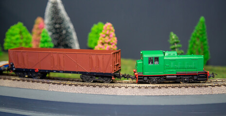 A mock-up of a green diesel electric locomotive with a freight car on a background of green fir trees. Toys electronics transport.