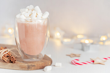 Sweet homemade hot chocolate or cocoa drink with marshmallow topping served in transparent glass cup or mug on wooden board with cinnamon sticks on white table with candy cane and blurred lights
