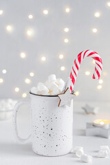 Homemade cocoa drink or hot chocolate with marshmallow sweets topping decorated with striped candy cane and star shaped decor served in cup or mug on white wooden table against blurred garland lights