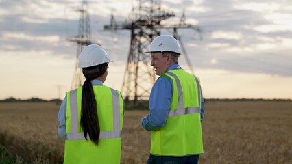 Happy electricians with tablet walk to power transmission lines at sunset countryside. Engineers with tablet discuss future of power plant technology by transmission lines. Electricians on field