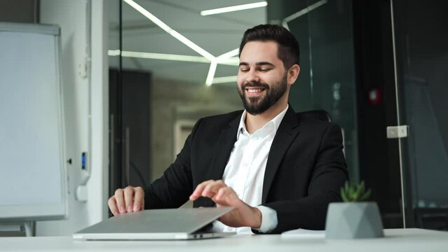 Positive caucasian businessman closing personal wireless laptop in end of intensive work day. Cheerful young bearded male using modern gadgets to solving job problems in spacious designer workplace.