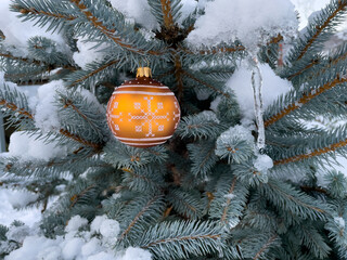 Christmas orange bauble with white ornament hanging outside on a snow covered blue spruce. Close up in a horizontal format. 