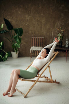 Beautiful young woman sitting on sunbad chair in a cozy living room and relaxing after workout or yoga. Healthy and sport concept