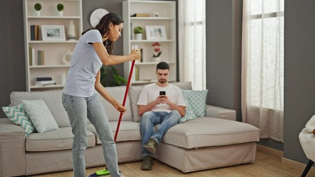 Beautiful couple expressing love, cleaning floor together at home. boyfriend casually texting on smartphone, standing indoors, shaking up household chores.