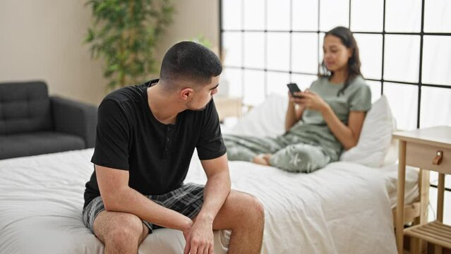 Beautiful couple disagreeing in their bedroom, sitting on the bed, expressing conflict while using a smartphone, a problem causing a serious expression