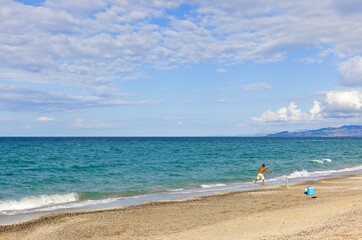 Fototapeta premium A Sardinian fisherman with a rod on Platamona Beach, Sardinia, Italy. The photo captures the beach, distant mountains, and beautiful clouds against the blue sky.