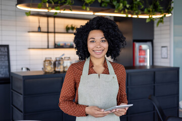 Portrait waitress of a small business owner of a cafe, woman in apron smiles and looks at camera with a tablet computer in her hands, an employee of a cafe of a restaurant near a coffee counter.