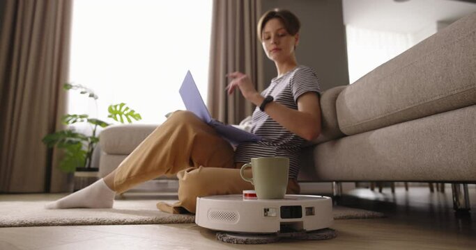 Female freelancer enjoyed peaceful day with her laptop in front of her. Robot vacuum cleaner brought her steaming cup of coffee. Beauty of smart technologies in comfort of her home. Cinematic AD