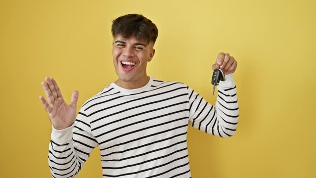 Joyful young hispanic man confidently holding car key, celebrating win with cheerful smile, ecstatic expression of achievement. handsome guy cheering victory over yellow isolated background