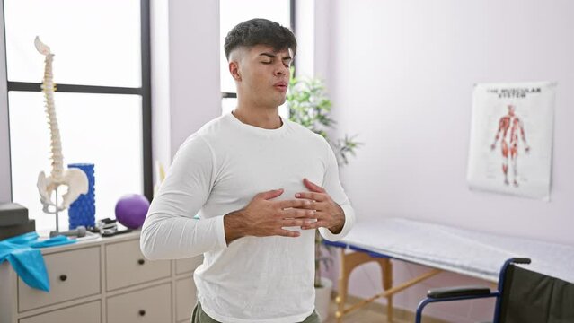 At his balance point, a portrait of a young, handsome hispanic man finding calm by meditating and taking a deep breath, relaxing in the indoor room of a rehab clinic, conquering respiratory problems