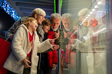 Two grandparents standing outside of a store window with their granddaughter and grandson in the city on a cold night, they are all wearing warm clothing and cheerfully chatting about gifts to buy.