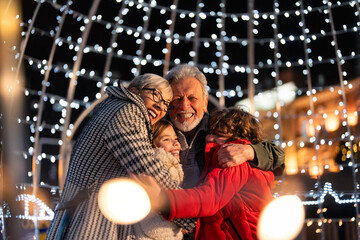 Loving grandparents and grandkids embracing at Christmas market in city center. Embraced seniors and children enjoying the Christmas fair during winter holidays. - obrazy, fototapety, plakaty