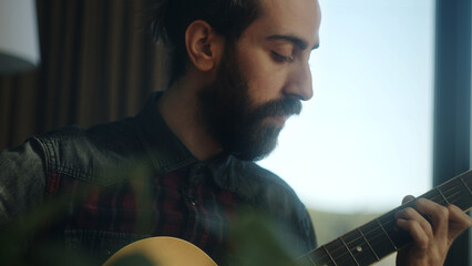 Bearded young adult man playing acoustic guitar while sitting on sofa at home. Camera moving down...