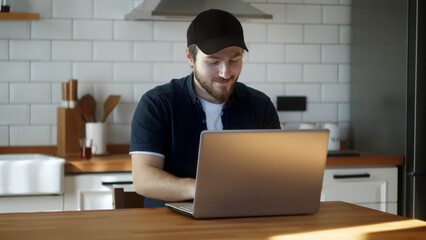 Happy young adult man in a hat sitting in the kitchen, chatting on his laptop with friends or family, doing some work