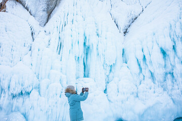 Female wonderer shooting video on smartphone of a big ice rock during winter adventure on Baikal lake