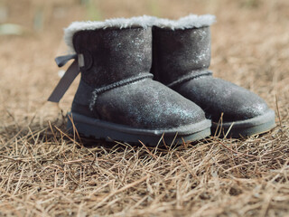 Horizontal close up side view of kids grey sheepskin boots with bows on a brown forest ground.