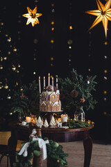 Christmas and New Year beautiful elegant family dinner table setting decorated with candles, fir tree branches, handmade craft toys. Gorgeous big homemade cake with gingerbread houses as centrepiece.