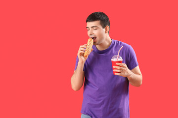 Young man with drink eating tasty french hot dog on red background