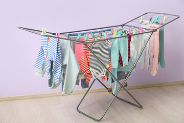 Clean baby clothes hanging on dryer near lilac wall