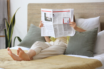Beautiful young woman reading newspaper in bedroom at home