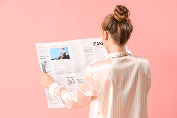 Young woman in pajamas reading newspaper on pink background