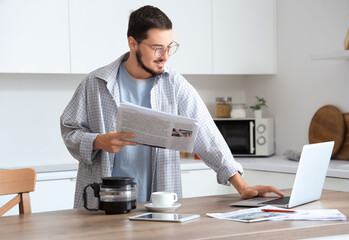 Young man with newspaper using laptop in kitchen - obrazy, fototapety, plakaty