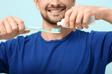 Handsome man squeezing tooth paste onto brush on blue background, closeup