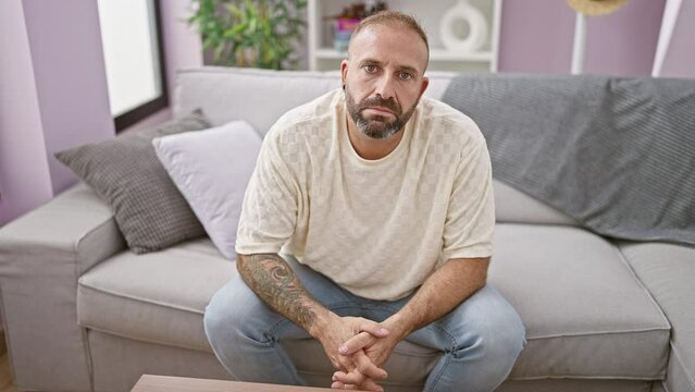 Handsome, worried young adult man sitting on sofa indoors pondering serious problems at home