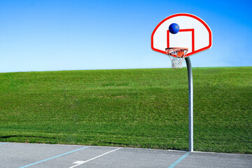 Blue basketball going up in the air over the hoop with rush green grass and blue sky background. Nobody at the court. Shooting shot of outdoor sport, challenge and achievement concept. Fun activity.