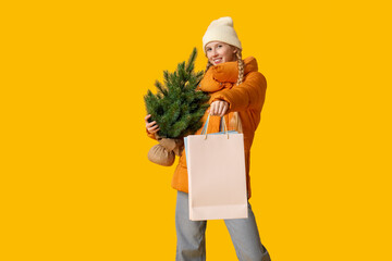 Young woman in warm winter clothes with shopping bag and Christmas tree on yellow background