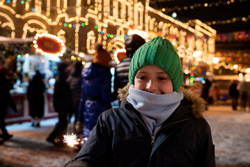 Obraz na płótnie Canvas happy boy in winter clothes holding a burning sparkler, having a joyful time at the Christmas market in the evening in the city center