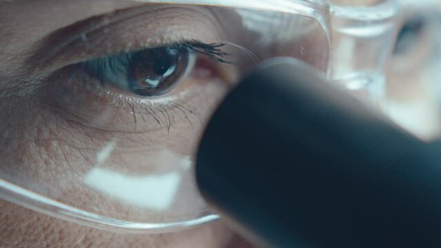 Brown eyes of female scientist in safety glasses looking through microscope oculars in laboratory