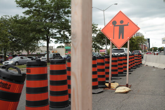 diamond orange construction sign with illustration of person holding up a stop sign in black print, road on left with traffic, wood post in foreground center frame