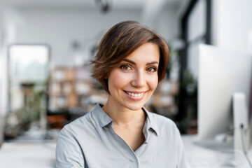 Portrait einer jungen Frau mit braunen Augen und kurzen dunklen Haaren die zuhause im Home Office Büro in die Kamera lächelt - obrazy, fototapety, plakaty