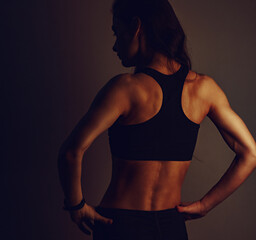 Female sport muscular woman posing in black sport bra showing the shoulders, blades and arms, standing on dark shadow background with empty copy space. Back view. Sporty - obrazy, fototapety, plakaty