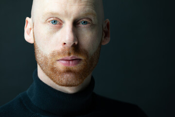 Mann mit Glatze,einem roten haarigen 3-Tage-Bart blickt selbstbewusst und stark mit seinen blauen Augen bekleidet mit einem Rollkragenpullover selbstsicher, vor einem schwarzen Hintergrund, in die cam