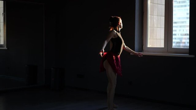 The ballerina performs several graceful spinning jumps in a row in a dark choreographic hall and stops to rest by the window