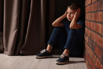 Fototapeta na wymiar Child abuse. Upset boy sitting on floor near brick wall indoors, space for text