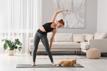 Beautiful woman with cute red cat practicing yoga on mat at home