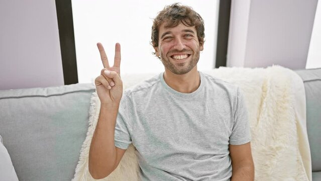 Confident young guy, a blond bearded winner, radiates happiness from his cozy sofa at home, sporting a victory sign, all smiles and winking for a friendly indoor portrait with a touch of comfort.