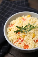 Tasty couscous with pepper, corn and arugula in bowl on wooden table, closeup