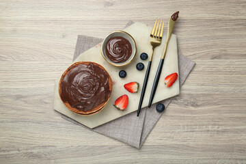 Delicious pancakes with chocolate paste, berries and cutlery on wooden table, top view