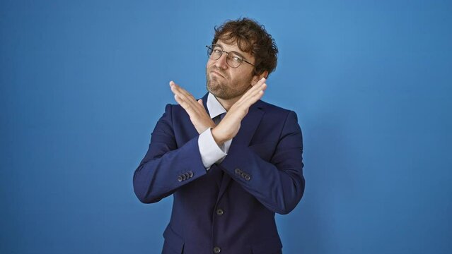 Angry young man in business suit expresses rejection with firm crossed arms gesture and negative sign, isolated on blue background