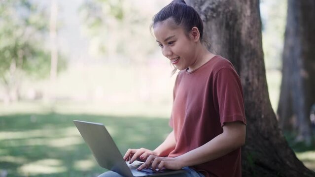 Asian female student is studying and researching course materials using her laptop under the tree in the garden, seeking a tranquil outdoor environment for learning and working outside.