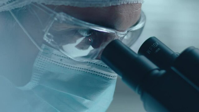 Close up shot of concentrated male scientist wearing face mask, safety glasses and medical hat looking through lab microscope