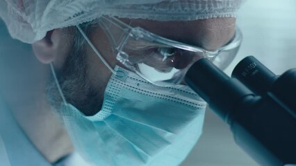 Close up view of focused chemist wearing face mask, safety glasses and medical hat looking through microscope and writing down results of lab research - Powered by Adobe